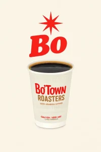 The image features a cup of Bo'Town Roasters coffee, with a rich black coffee visible at the top. The cup is white with a design that includes "Bo'Town Roasters" in bold red and brown letters, along with a cautionary note at the bottom. Above the cup, there is bold red "Bo" lettering and a starburst graphic. The background is a soft cream color, highlighting the cup and the coffee.
Bojangles Menu