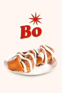 "A Cinnamon twistbiscuit featuring a warm, soft biscuit topped with sweet glaze and filled with cream all drizzled with white icing. The biscuit is placed on a white plate with the word 'Bo' in bold red letters and a starburst above it, set against a plain light-colored background.Bojangles Menu